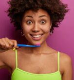 Head shot of attractive curly haired woman has toothy healthy smile, holds toothbrush, cleans teeth, has wide opened eyes, wears casual vest, isolated over purple background, has perfect smooth skin