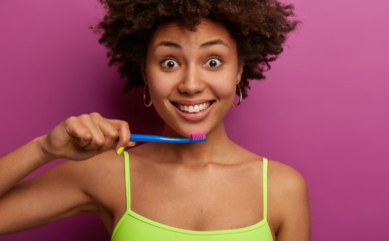 Head shot of attractive curly haired woman has toothy healthy smile, holds toothbrush, cleans teeth, has wide opened eyes, wears casual vest, isolated over purple background, has perfect smooth skin
